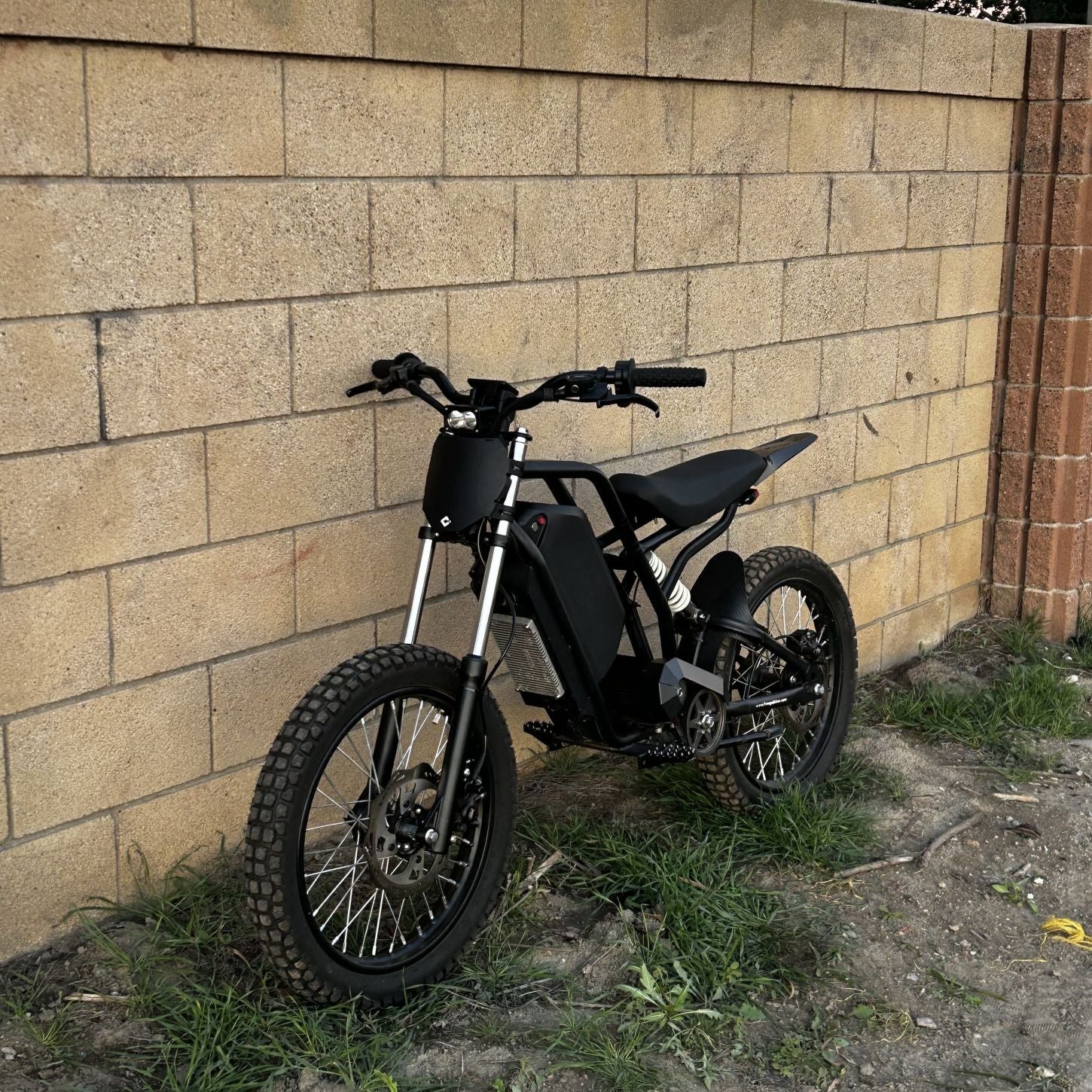 Freego Nova 3 electric dirt bike parked on grass in front of a tan brick wall, featuring knobby off-road tires, front suspension fork, rear shock absorber, and a minimalist black frame with centrally housed motor and battery.