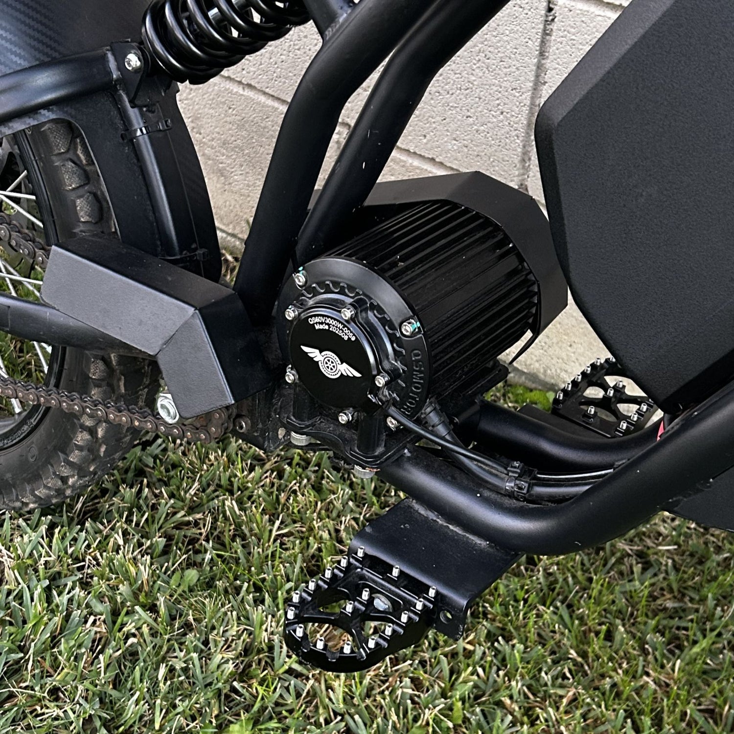 Close-up of the Freego Nova 3 electric dirt bike’s motor and drivetrain, showing a QS MOTOR 3000W cylindrical motor, chain drive, rear suspension spring, and rugged foot pedals mounted on a black frame.