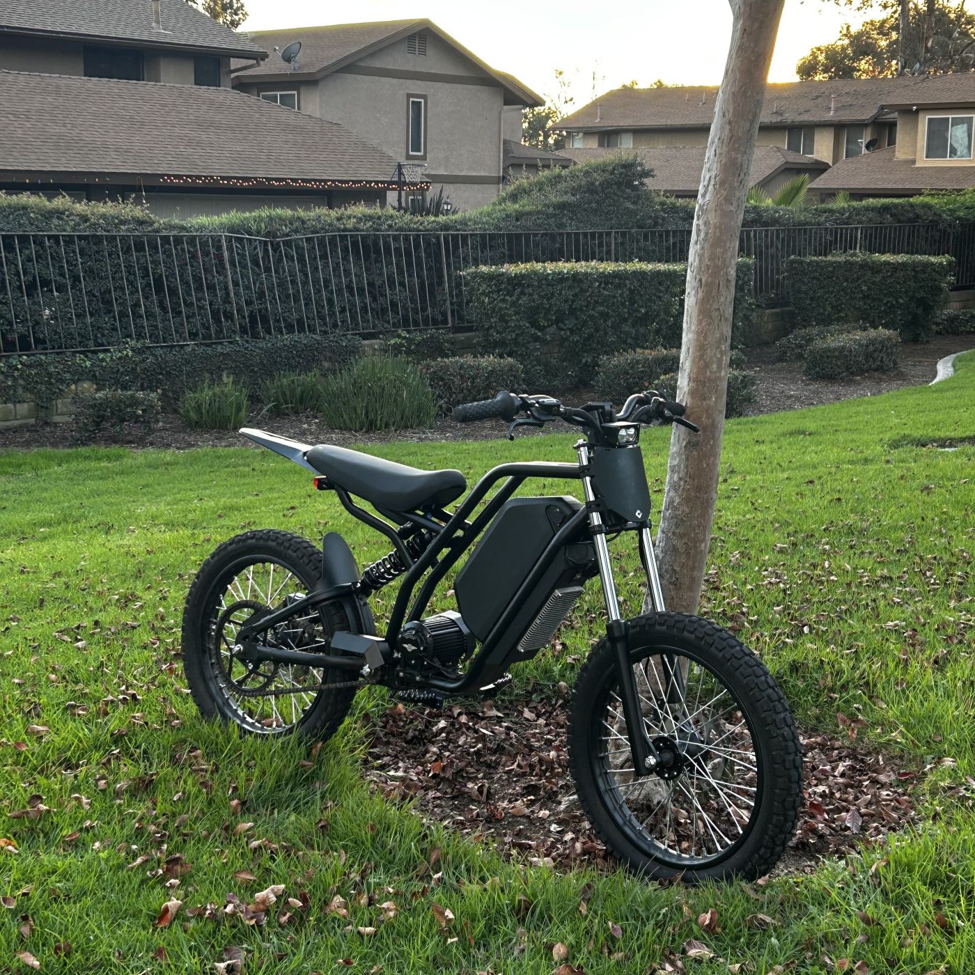 Freego Nova 3 electric dirt bike parked on grass near a tree in a residential area, featuring a black frame, knobby tires, front suspension fork, and centrally mounted battery pack.