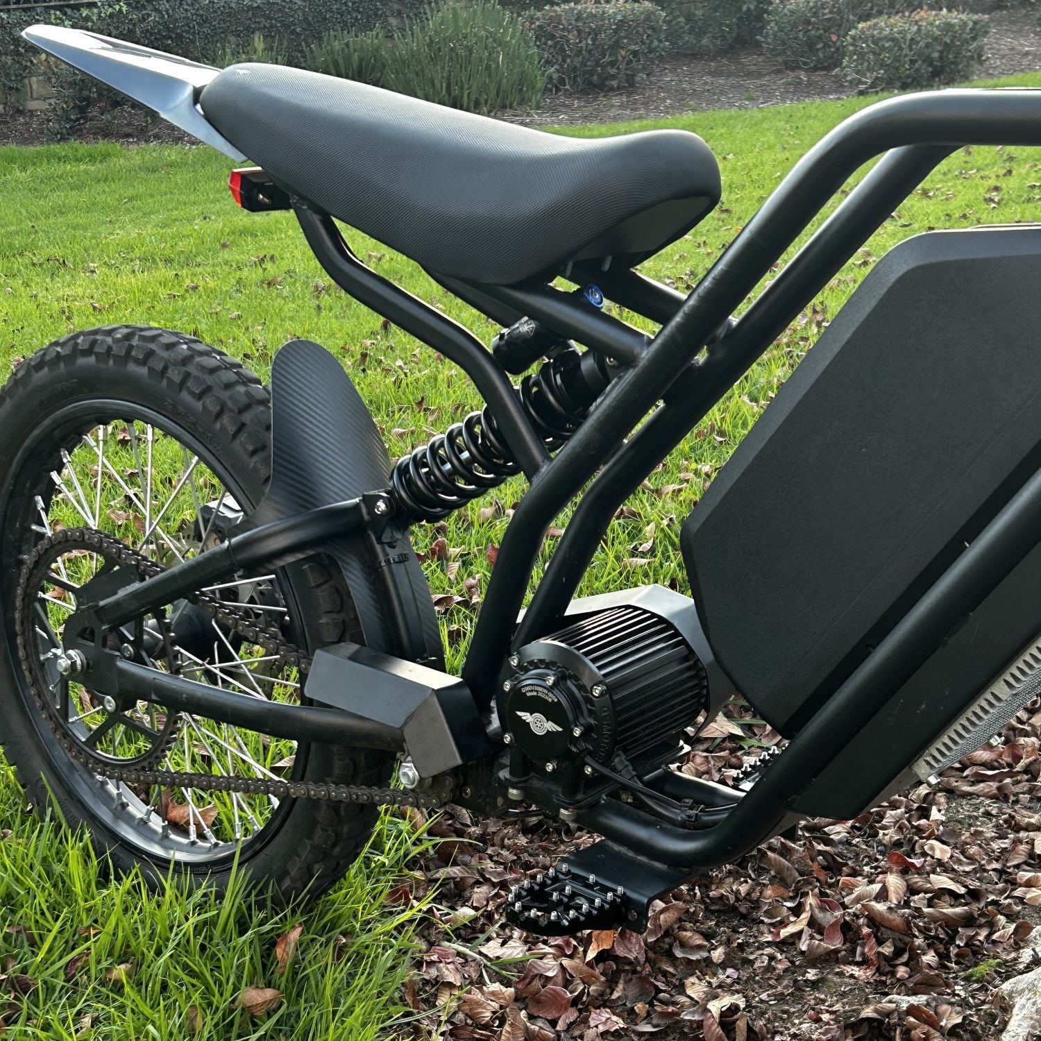 Close-up of the rear section of the Freego Nova 3 electric dirt bike showing the knobby tire, coil spring rear suspension, chain drive, foot peg, and cylindrical motor mounted low on the black tubular frame, with a large battery pack integrated above.